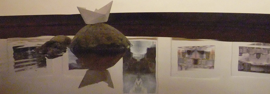 Fotokunst spiegelt sich im Wasser des Objektes Wohnzimmer-Weltmeer. unterbrochen wird die Spiegelung von einem Stin, auf dem ein gefaltetes Papierboot steht.