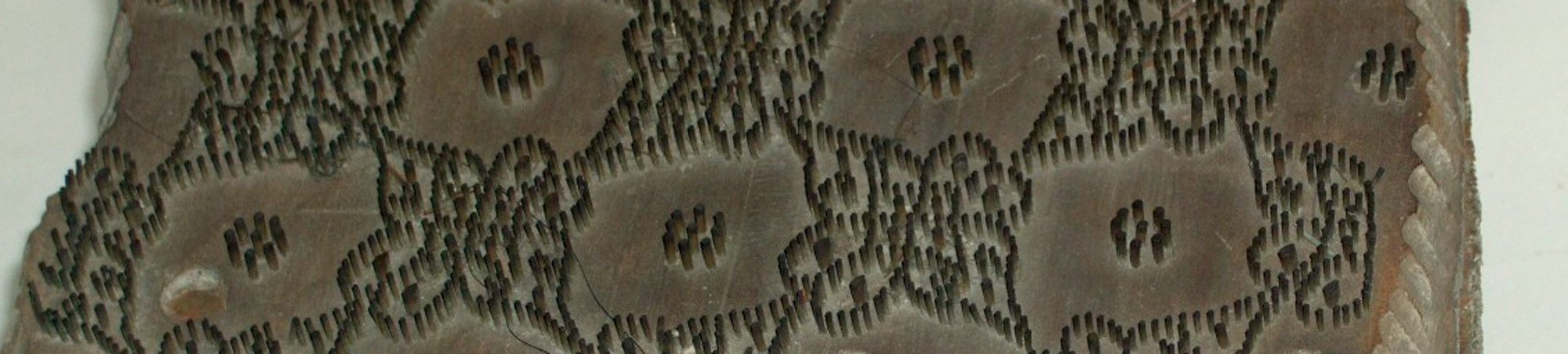 Fast quadratischer Druckstock, bestehend aus zwei aufeinander genagelten Holzbrettern (unten Weich- oben Hartholz) An der Unterseite Handgriff eingeschnitzt. Auf der Oberseite mit Metallstiften beschlagen, die ein miteinander verbundenes Muster aus gleichen Blüten bilden. Picotmodel.