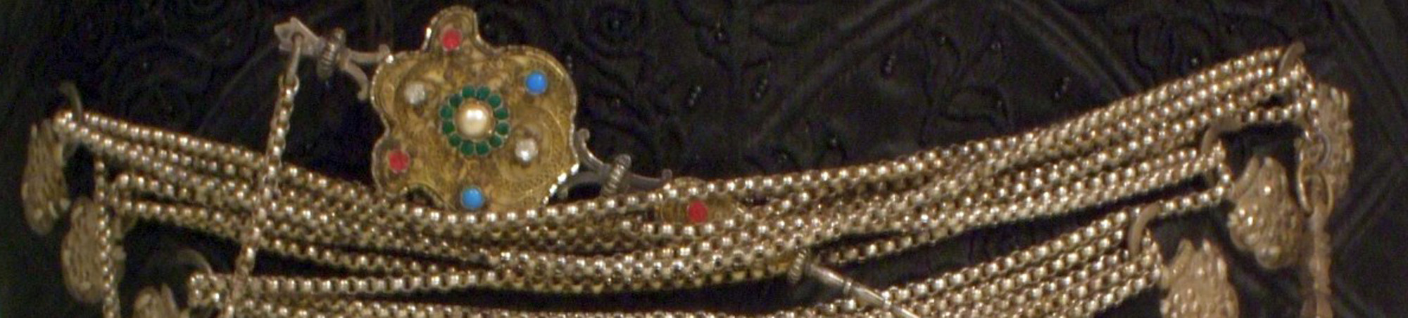 Mieder aus schwarzer Seide. Reich mit schwarzem Garn ornamental bestickt. Dazwischen schwarze Glasperlen. Innen mit rotgestreiftem, weißen Leinen gefüttert. Auf der Vorderseite links und rechts je sechs silberfarbene Geschnürhaken. 