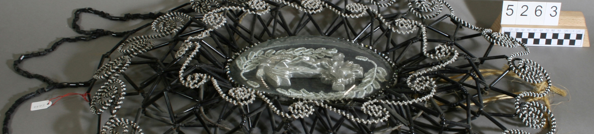 Grabschmuck ovale Kartusche, inliegend Kruzifix (silberfarbenes Gußmetall), weiß-opake Flechtgirlanden, außen geflochtene Girlanden auf Hohlkonstruktion.