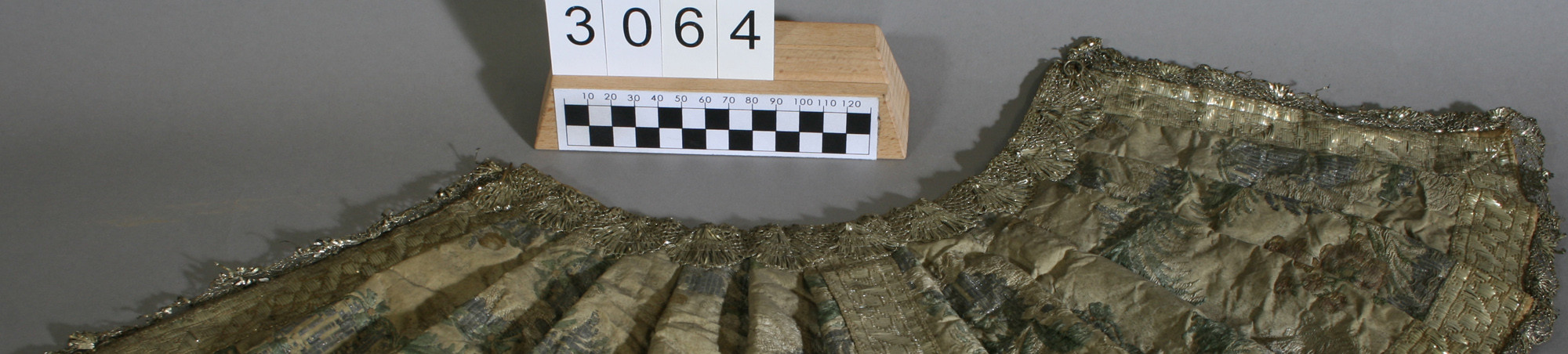 Mäntelchen aus ursprünglich hellem Seidenstoff mit eingewebten bunten Blumen. Gesäumt und in der Mitte mit einer goldfarbenen Metallborte verziert. An allen vier Kanten eine Metallklöppelspitze angesetzt. Helles Baumwollfutter mit Zwischenlage aus Papier.