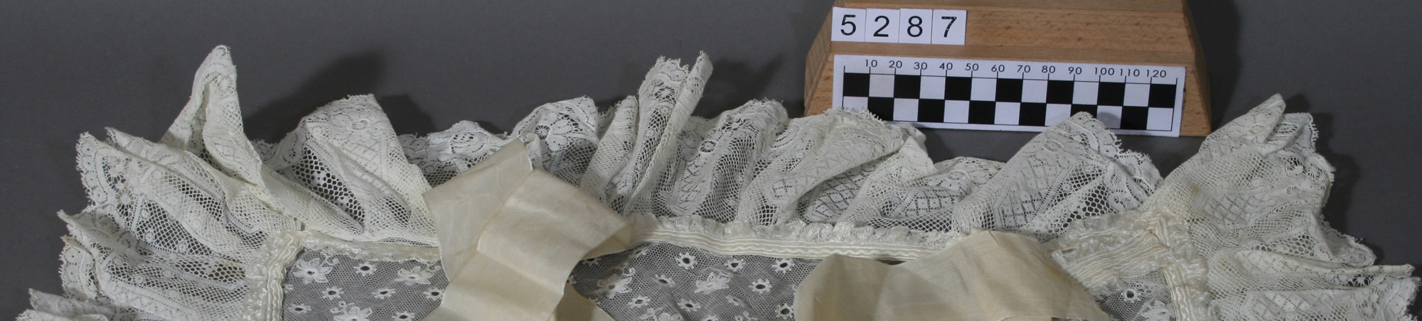 Decke Teil einer Taufbekleidung, aus cremefarbigem besticktem Tüll mit floraler Lochstickerei, diagonal aufgenähte Seidenborte, umlaufend breite Spitzenborte, beidseits je zwei Bindebänder aus cremefarbigem Seidenmoireè. 