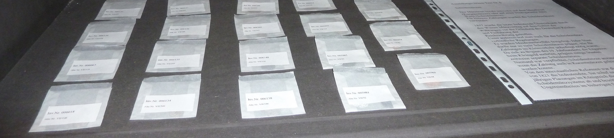 Inventarbeutelchen mit Münzen neben dem geplanten Begleittext auf schwarzem Vitrinenboden.