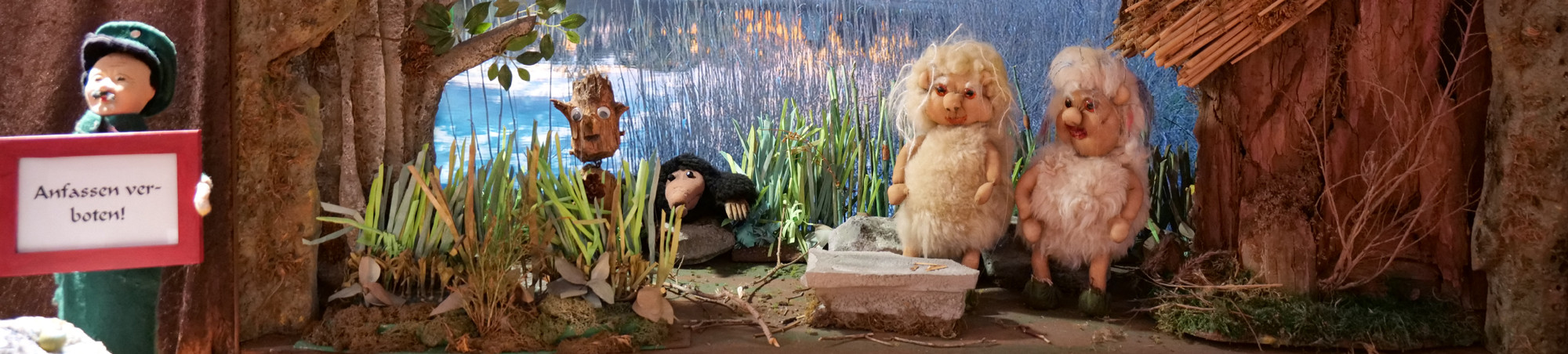 Vor einer Seehütte sitzen zwei Moosmännchen. In der Uferzone sind ein Maulwurf und ein Wurzelmännchen unterwegs.