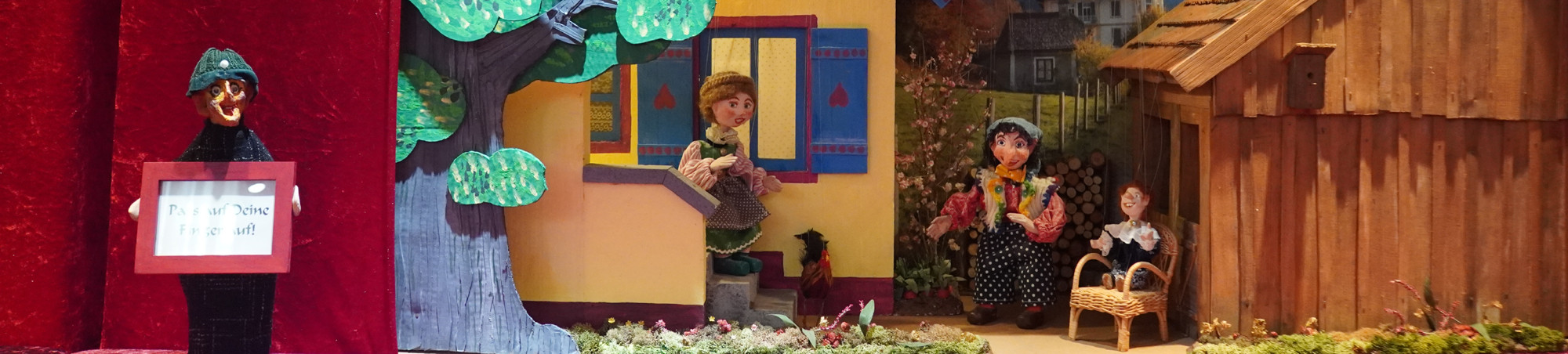 Kasperl, seine Frau und sein Kind sind im Garten zu Gange. Rechts steht ein Schuppen, links das Haus und ein großer Baum. Der Zwischenraum gibt den Blick auf den Weg, eine Nachbarvilla und Landschaft frei.