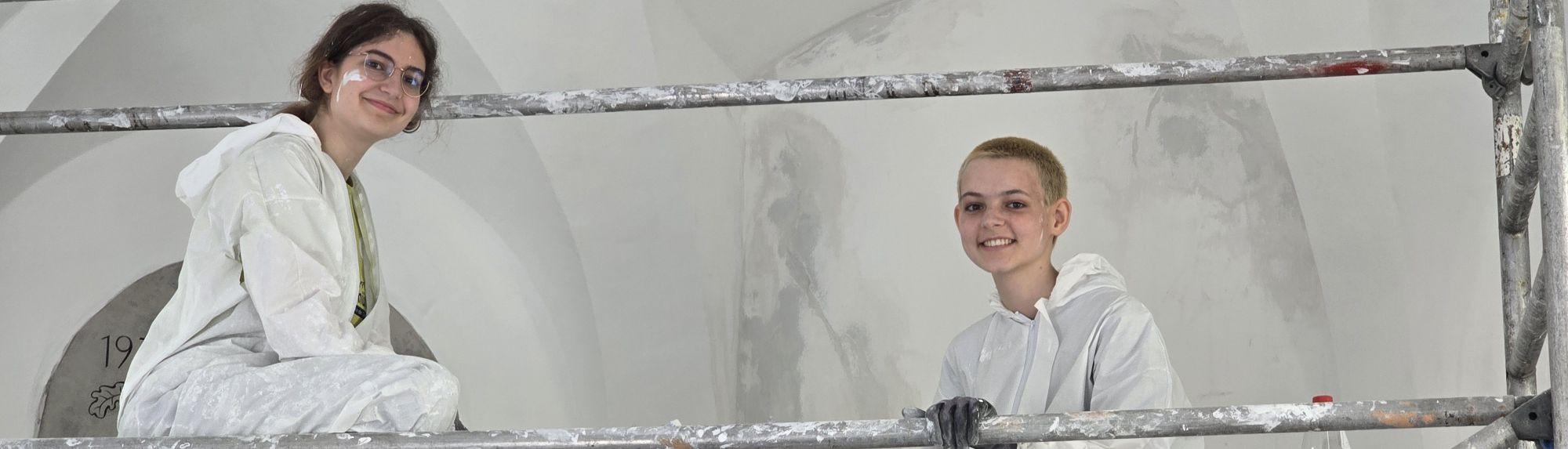 Zwei M&auml;dchen auf einem Ger&uuml;st unter einem Gew&ouml;lbe. Sie streichen die Wand mit wei&szlig;er Farbe.